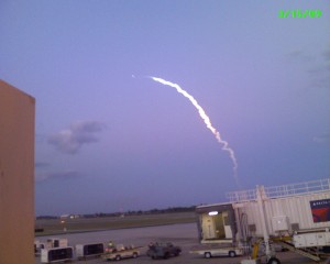 Trail from the Space Shuttle Discovery out the portside window of an airplane.